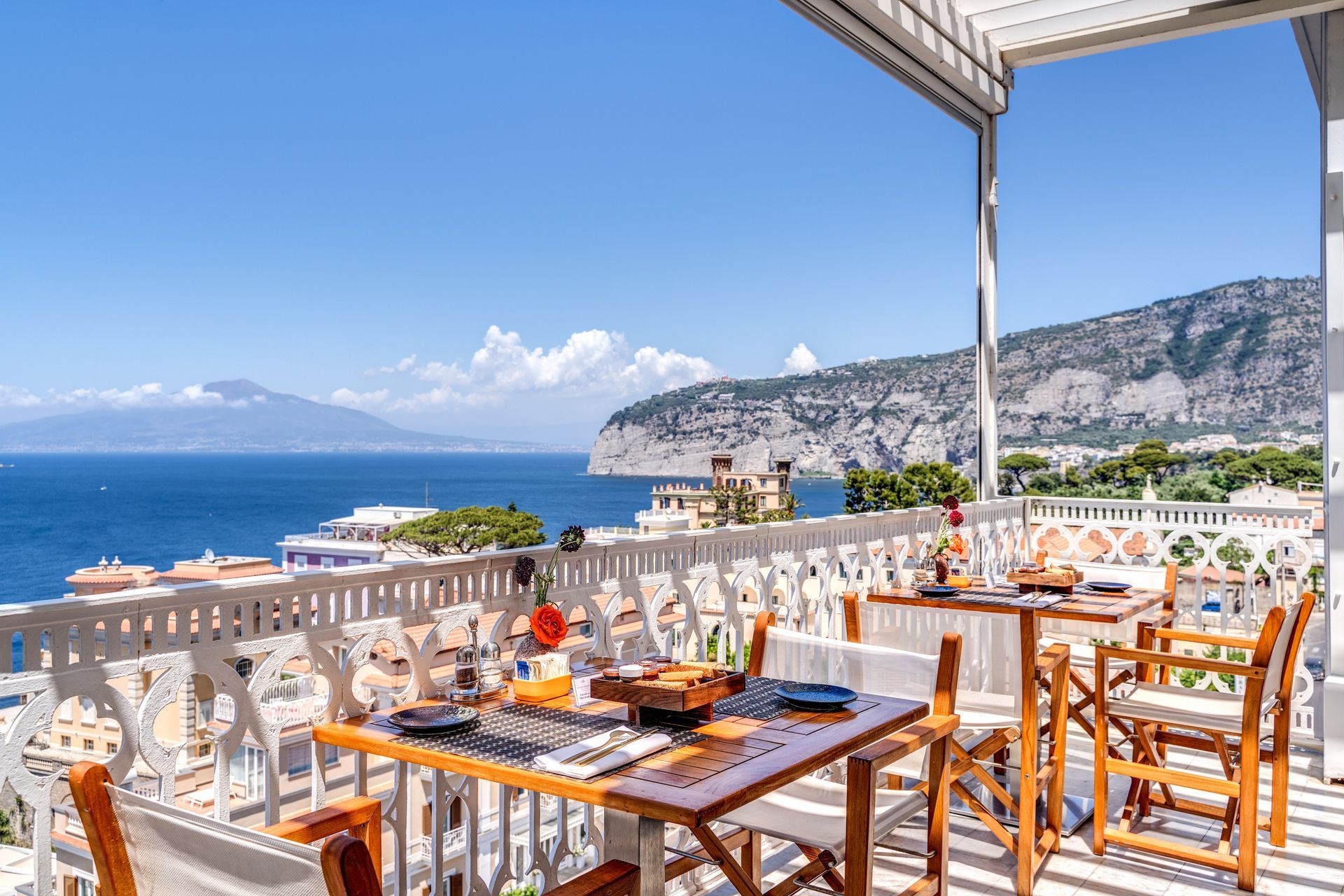 An outdoor balcony restaurant with wooden tables and chairs overlooking a sunny Mediterranean coastline and sea.