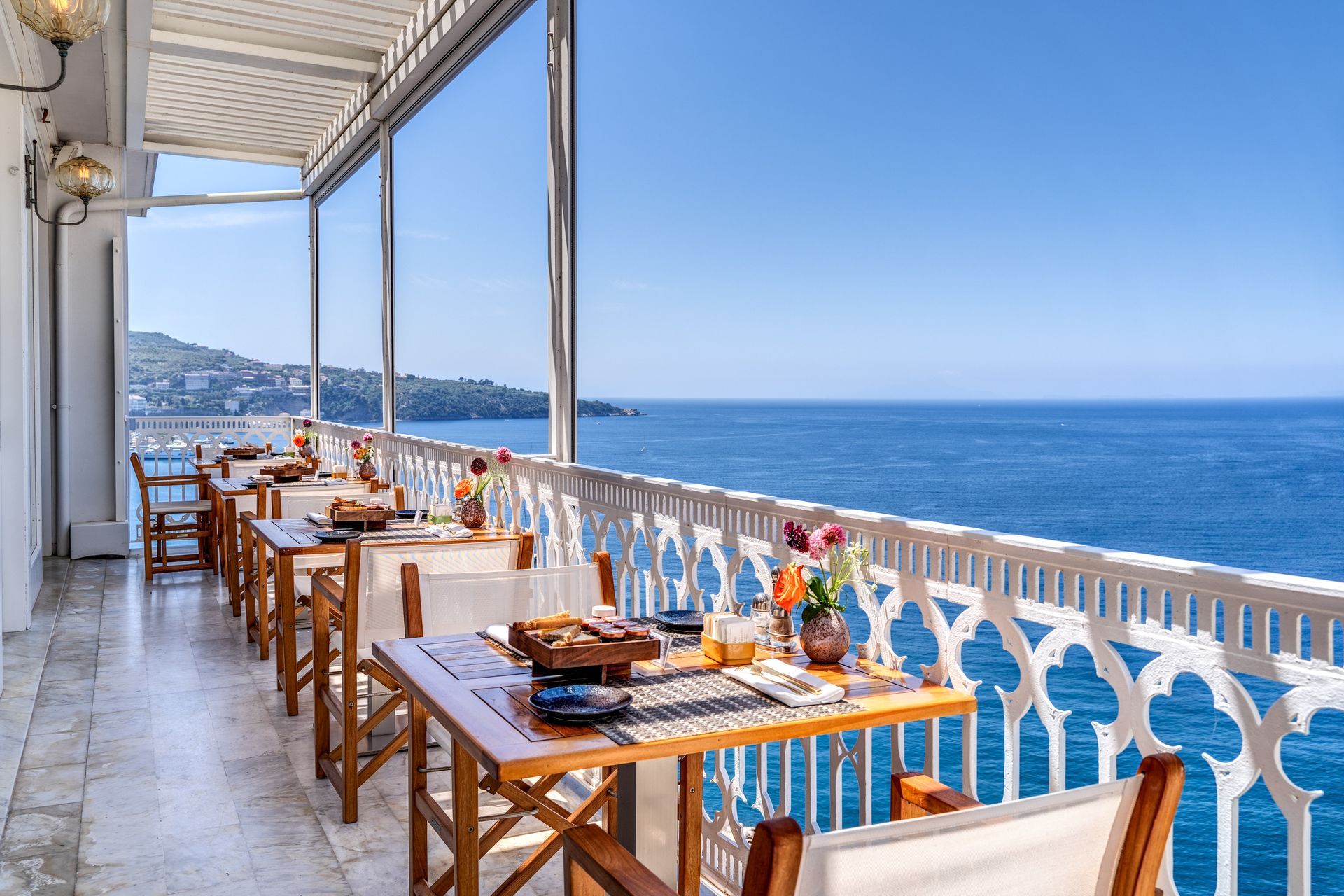 Outdoor patio restaurant terrace overlooking a deep blue sea with white wooden railings and tables set for breakfast.