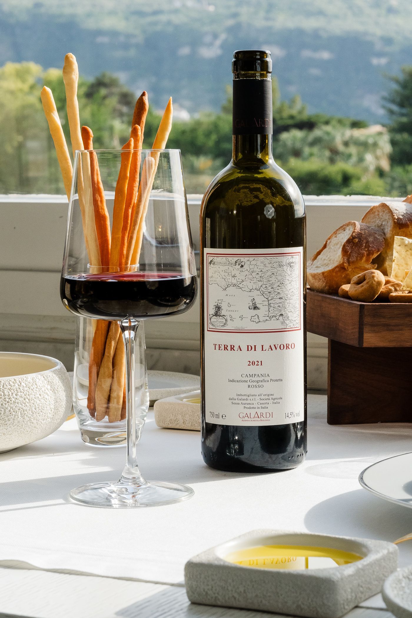 Una bottiglia di vino rosso, un bicchiere pieno di grissini e una ciotolina d'olio su un tavolo all'aperto, sotto il sole.