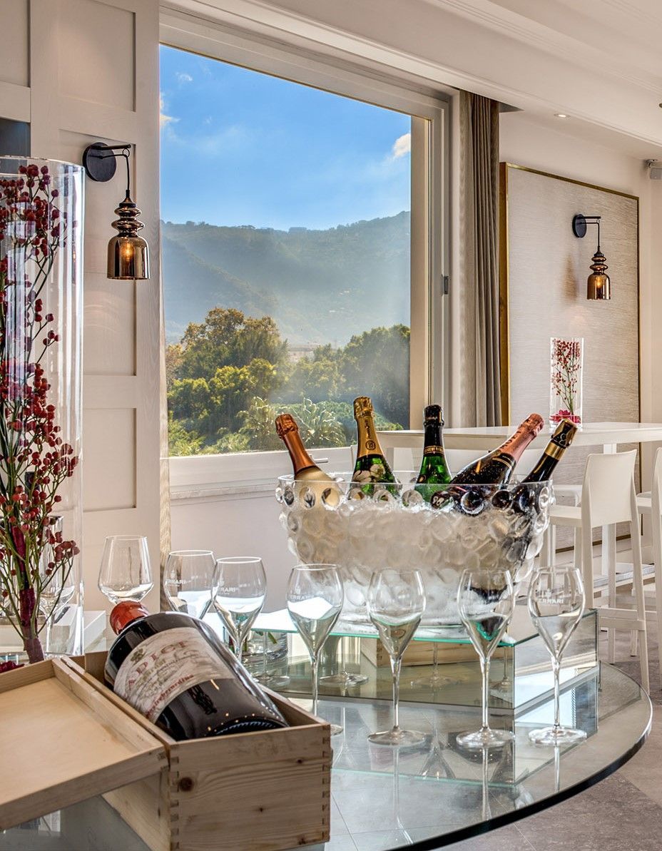 Champagne bottles in an ice bucket and a wine bottle in a wooden crate on a glass table before a window with a mountain view.