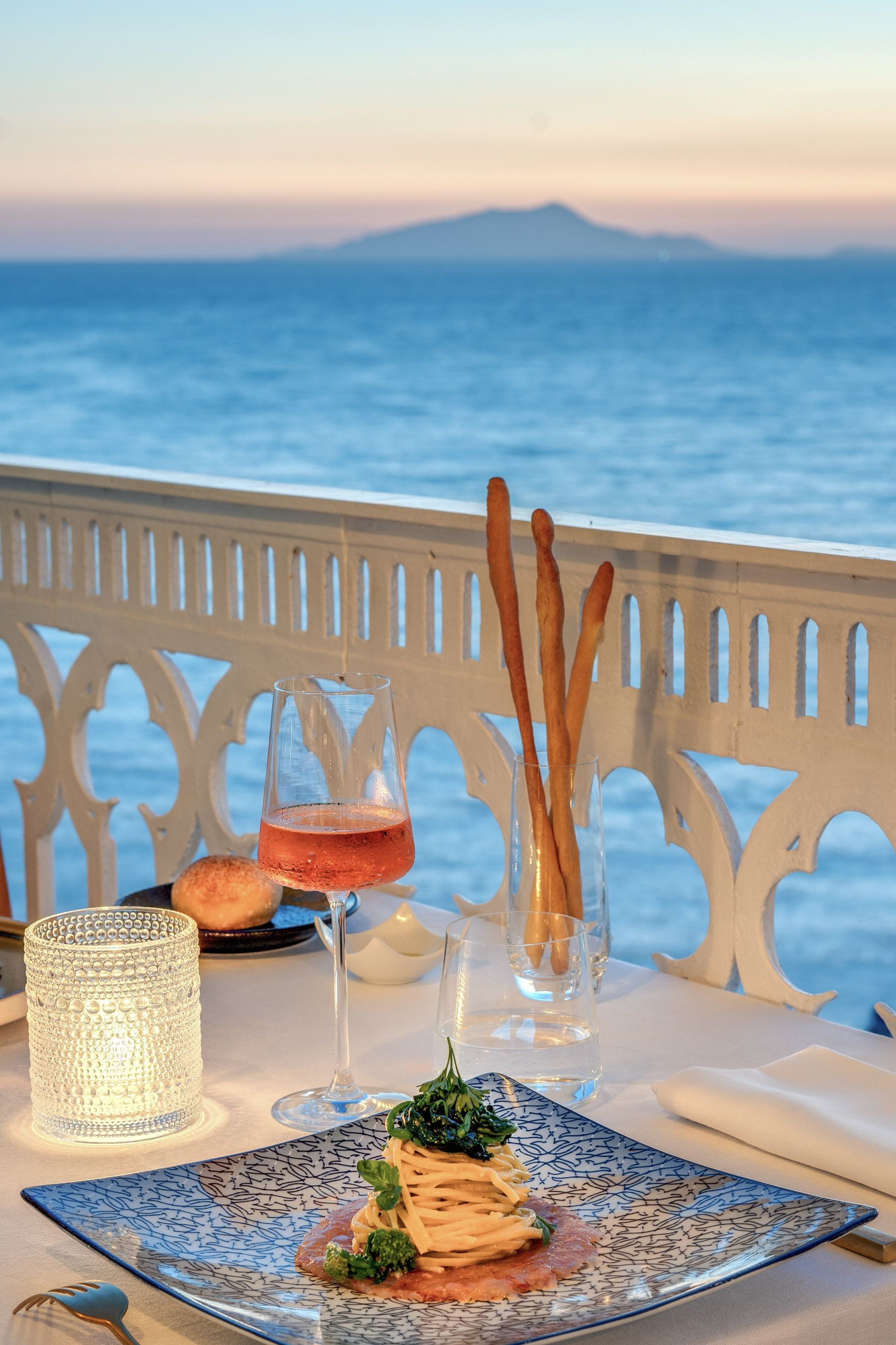 A plate of food and a glass of rosé on a table overlooking the sea at sunset, framed by a white ornate railing.