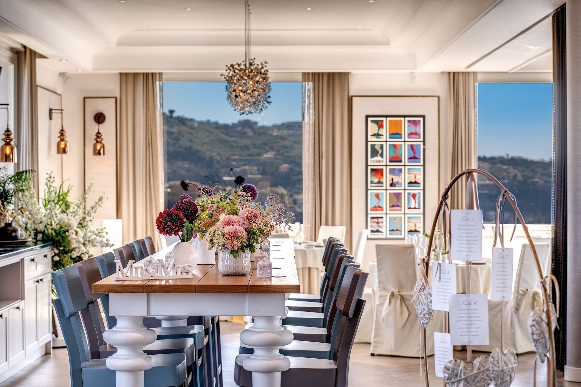 A sunlit dining room with a long table, blue chairs, floral centerpieces, and large windows overlooking a green landscape.