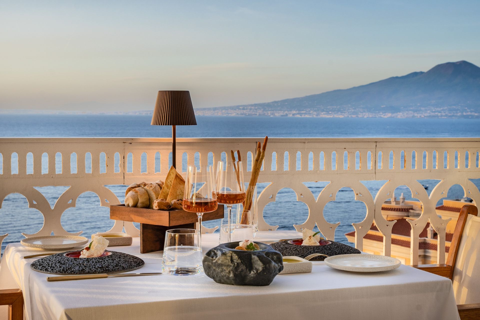 A set table on a terrace overlooking the sea and a distant mountain, featuring minimalist tableware and an ambient lamp.