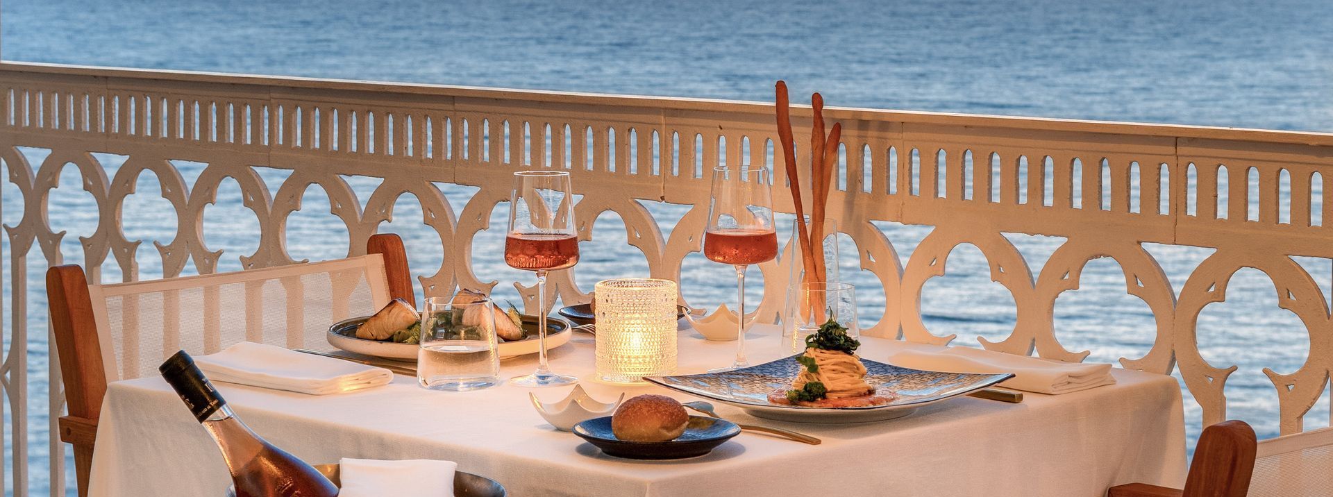 A table set for two on a balcony overlooking the water at sunset, featuring wine, plates, and candles.