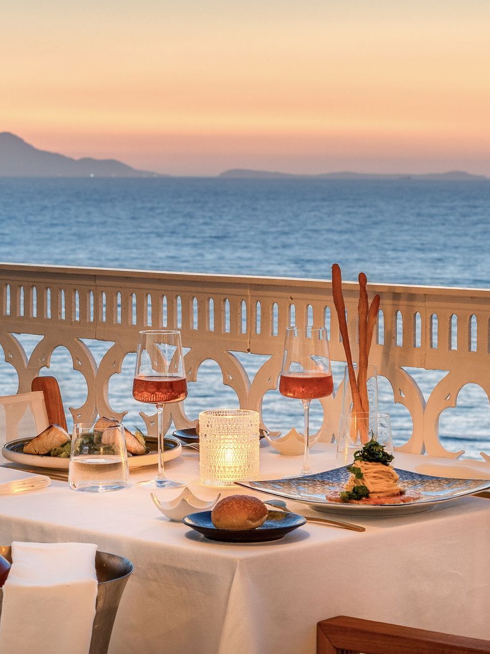 Un tavolo per due apparecchiato per una cena su un balcone con vista sul mare al tramonto, con vino e piatti di cibo.