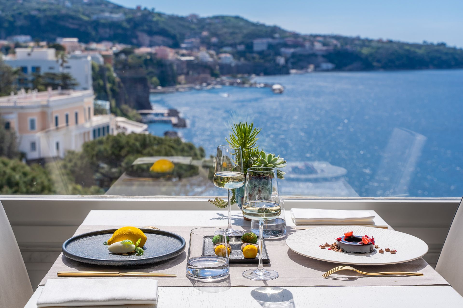 A table set for a meal with plates of food and wine glasses, overlooking a coastal town and the sea from a balcony.