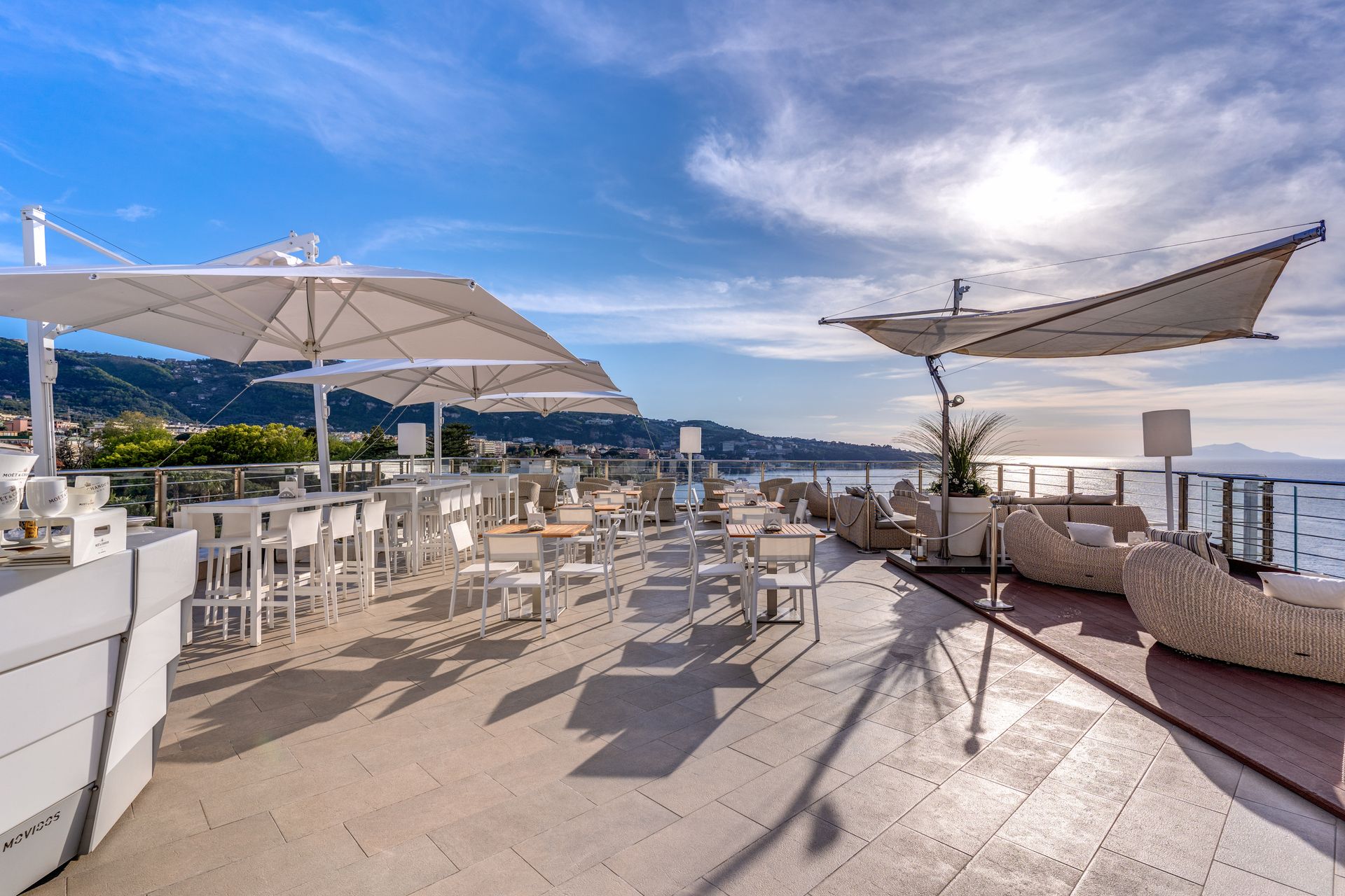 Un patio soleggiato con vista sull'oceano, arredato con tavoli, sedie, ombrelloni bianchi e comodi divani in vimini.