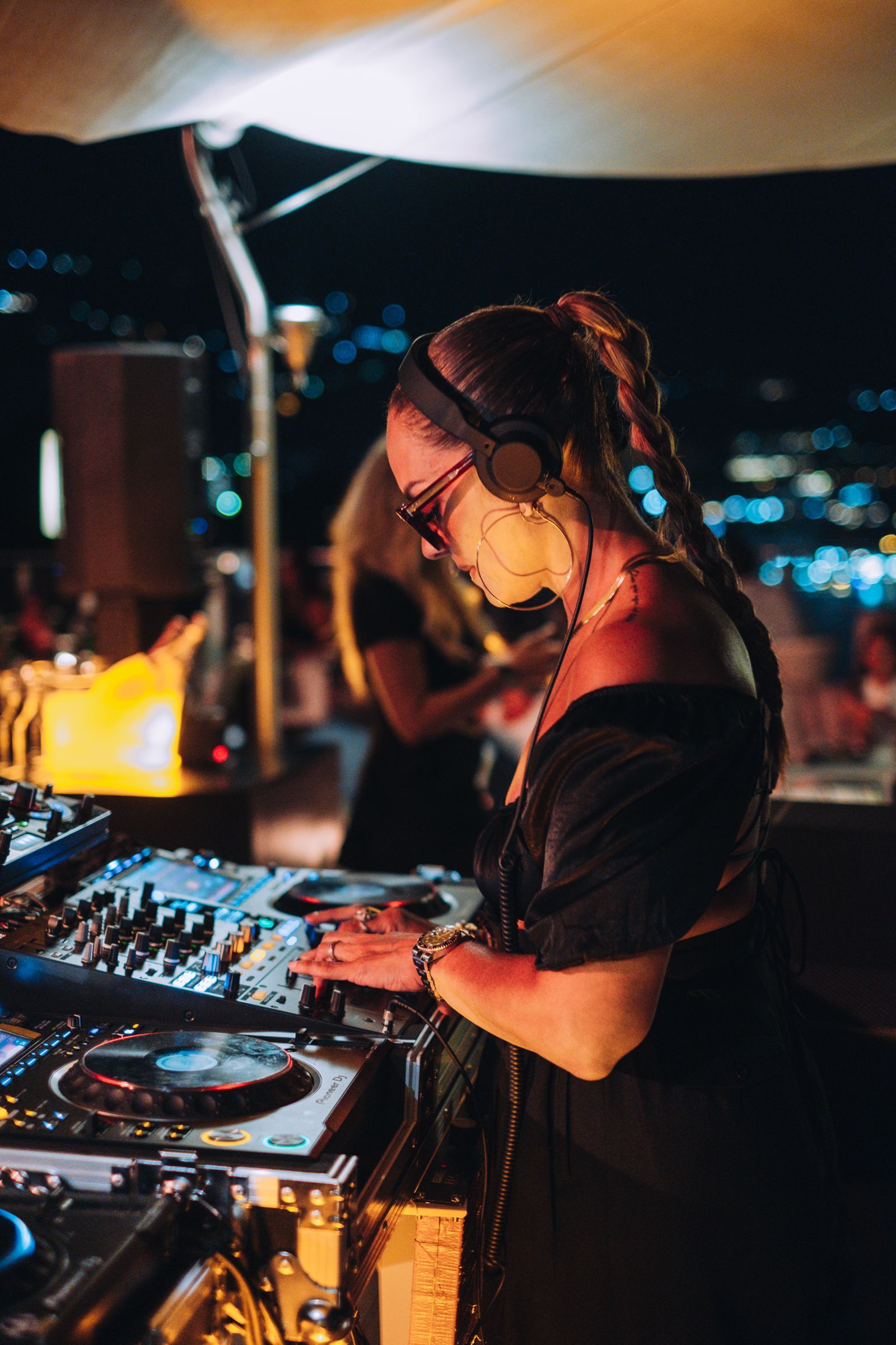 Una persona con i capelli intrecciati, che indossa cuffie e occhiali da sole, fa la DJ a un evento notturno all'aperto, con le luci della città sullo sfondo.