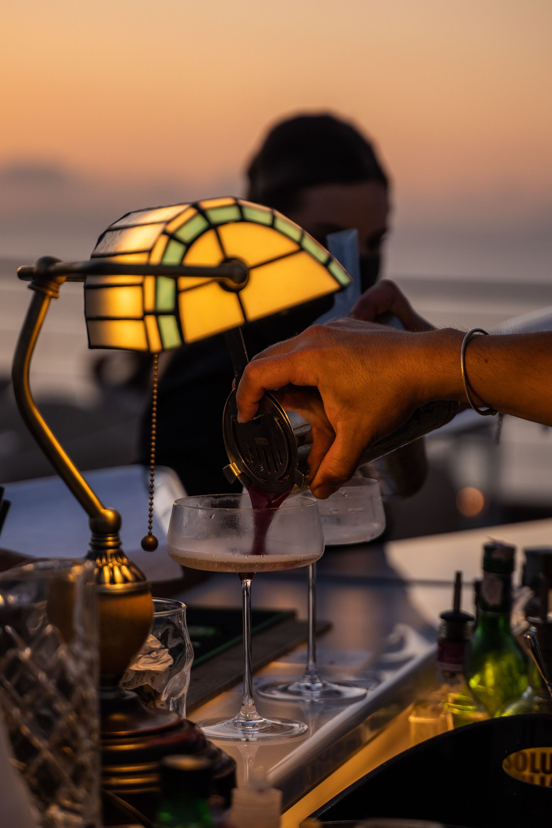 Un barista versa un cocktail scuro in un bicchiere sul bancone del bar, illuminato dalla calda luce di una lampada da banca d'epoca al tramonto.
