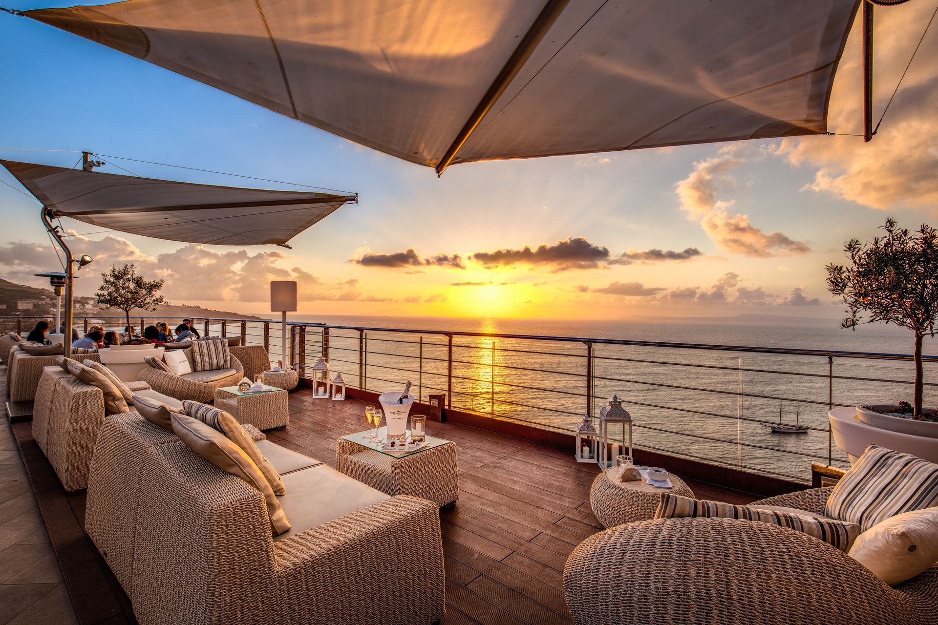 An outdoor terrace with wicker seating overlooks a calm sea at sunset under large, geometric shade sails.