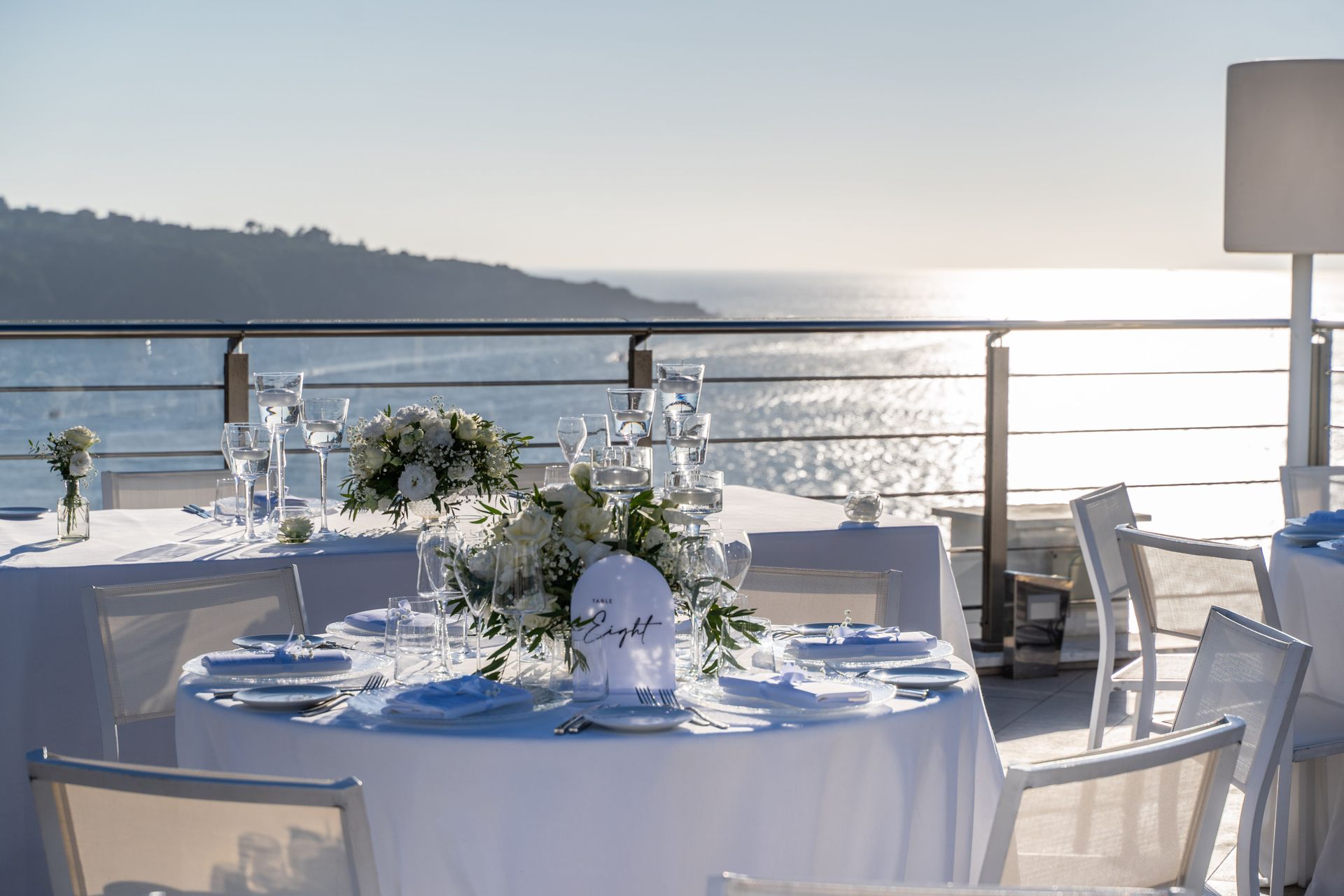 Round tables with white linens and floral centerpieces set on an outdoor terrace overlooking the sea at sunset.