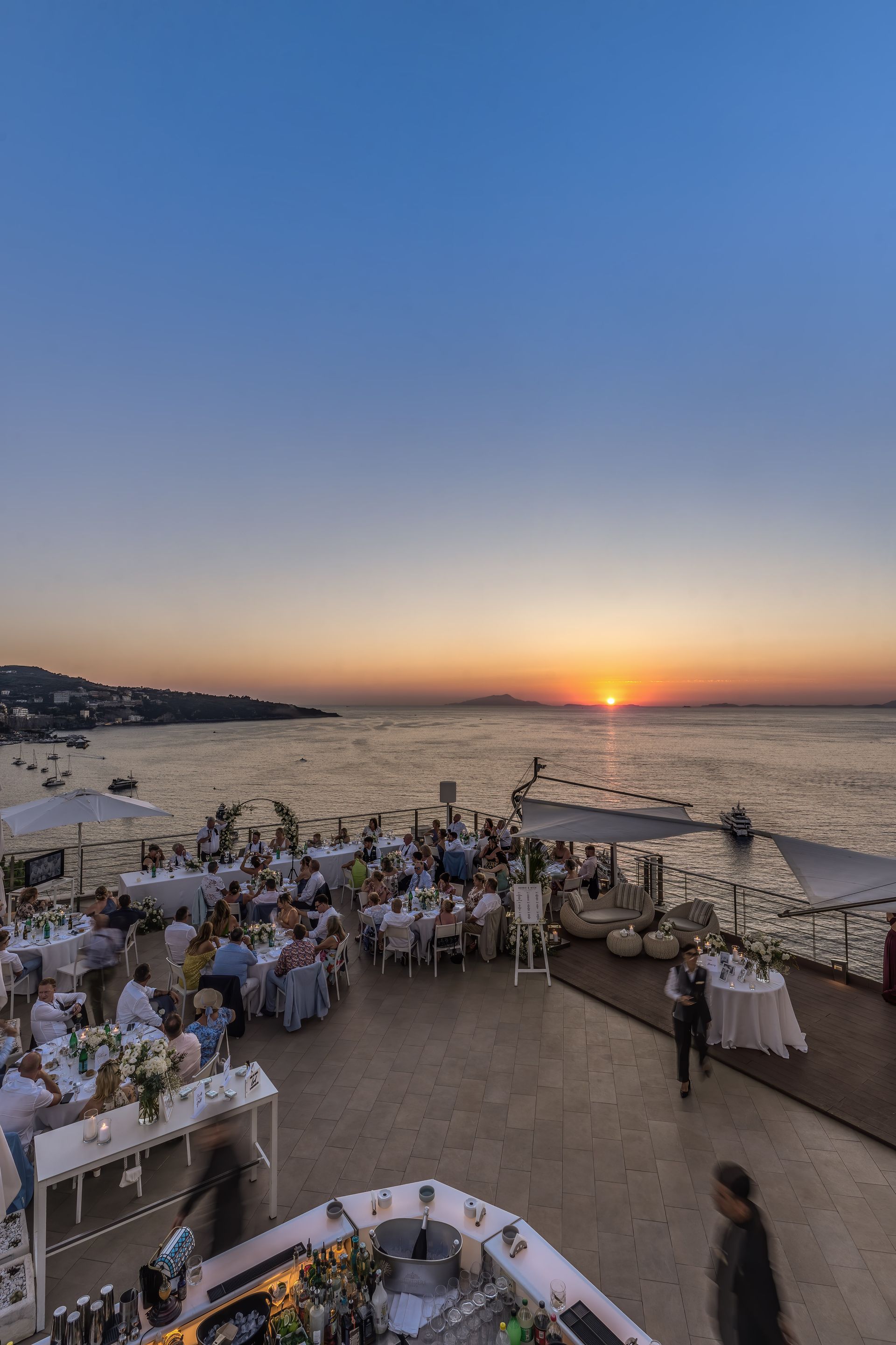 An outdoor dinner event at a coastal venue during a sunset with an orange sun over the sea and guests at white tables.