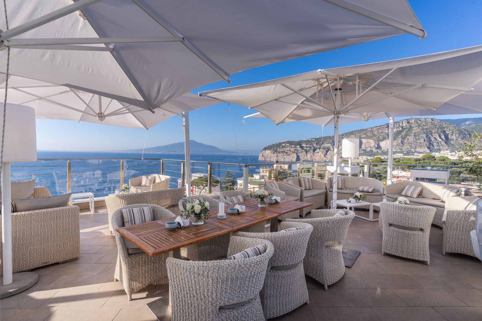 Outdoor terrace seating with wicker furniture under large white umbrellas, overlooking the sea and a coastal mountain.