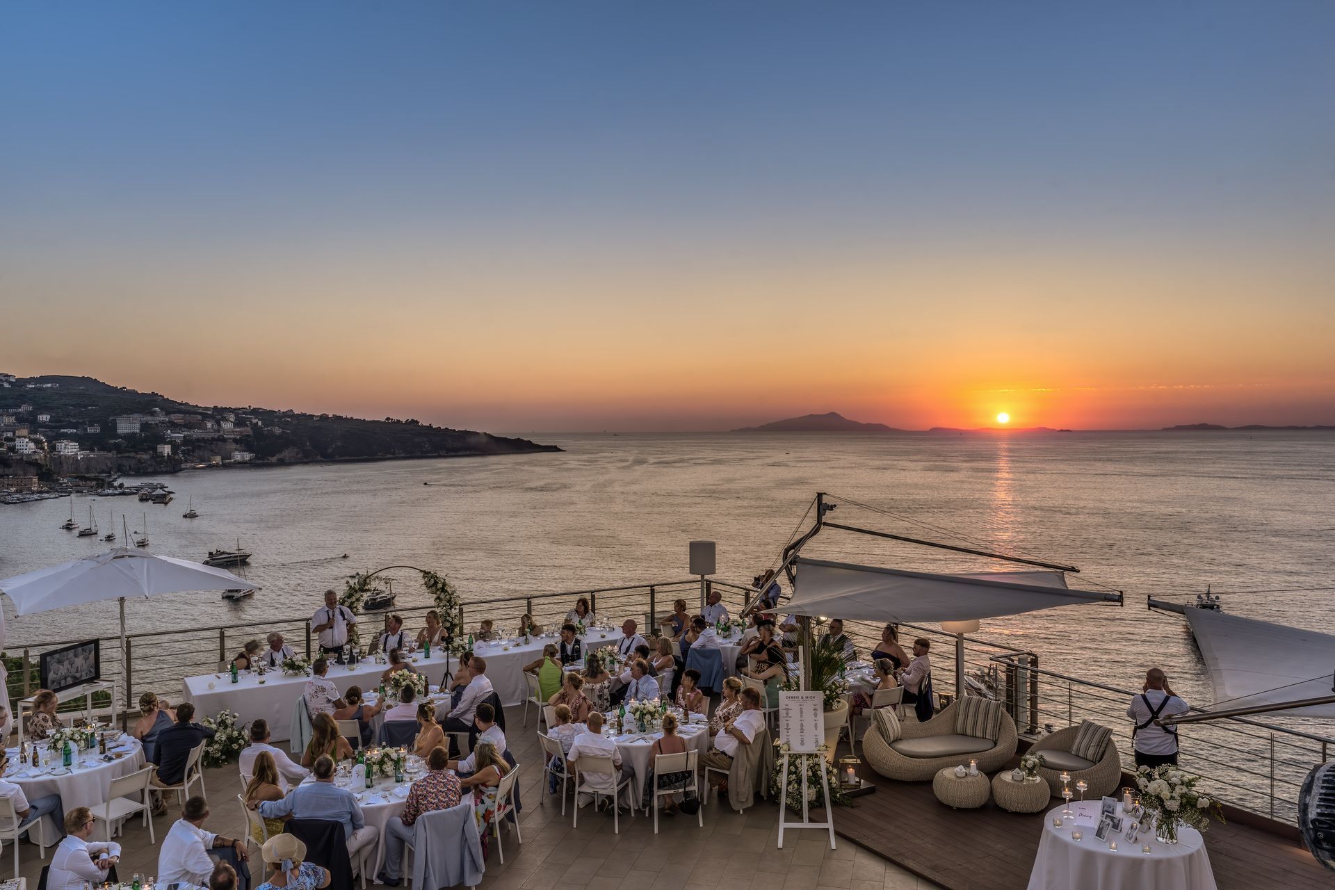 A coastal terrace with dining tables set for guests at sunset, overlooking a calm sea with distant land on the horizon.