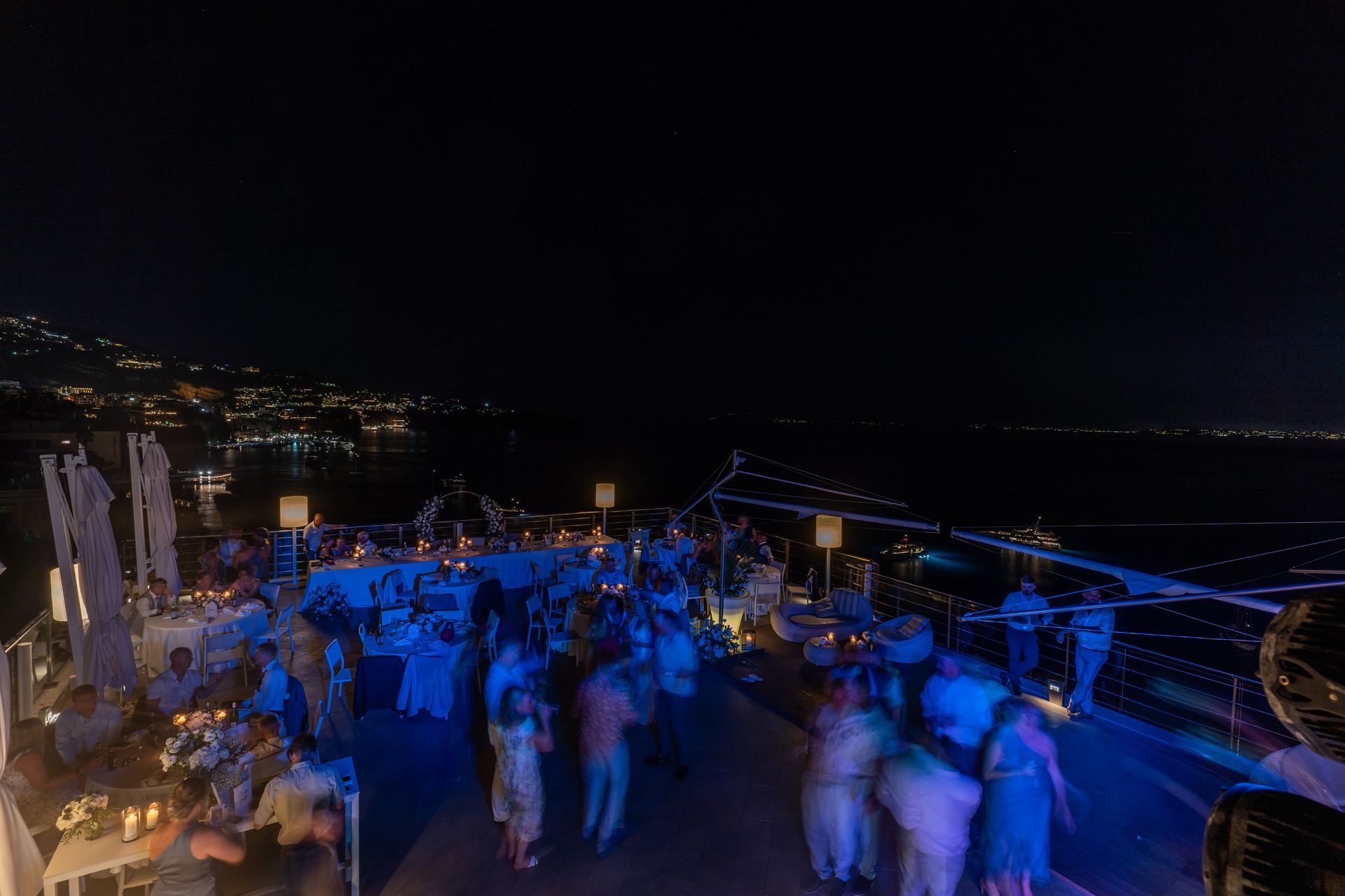 An outdoor night party on a balcony overlooking the sea, with guests socializing under string lights.