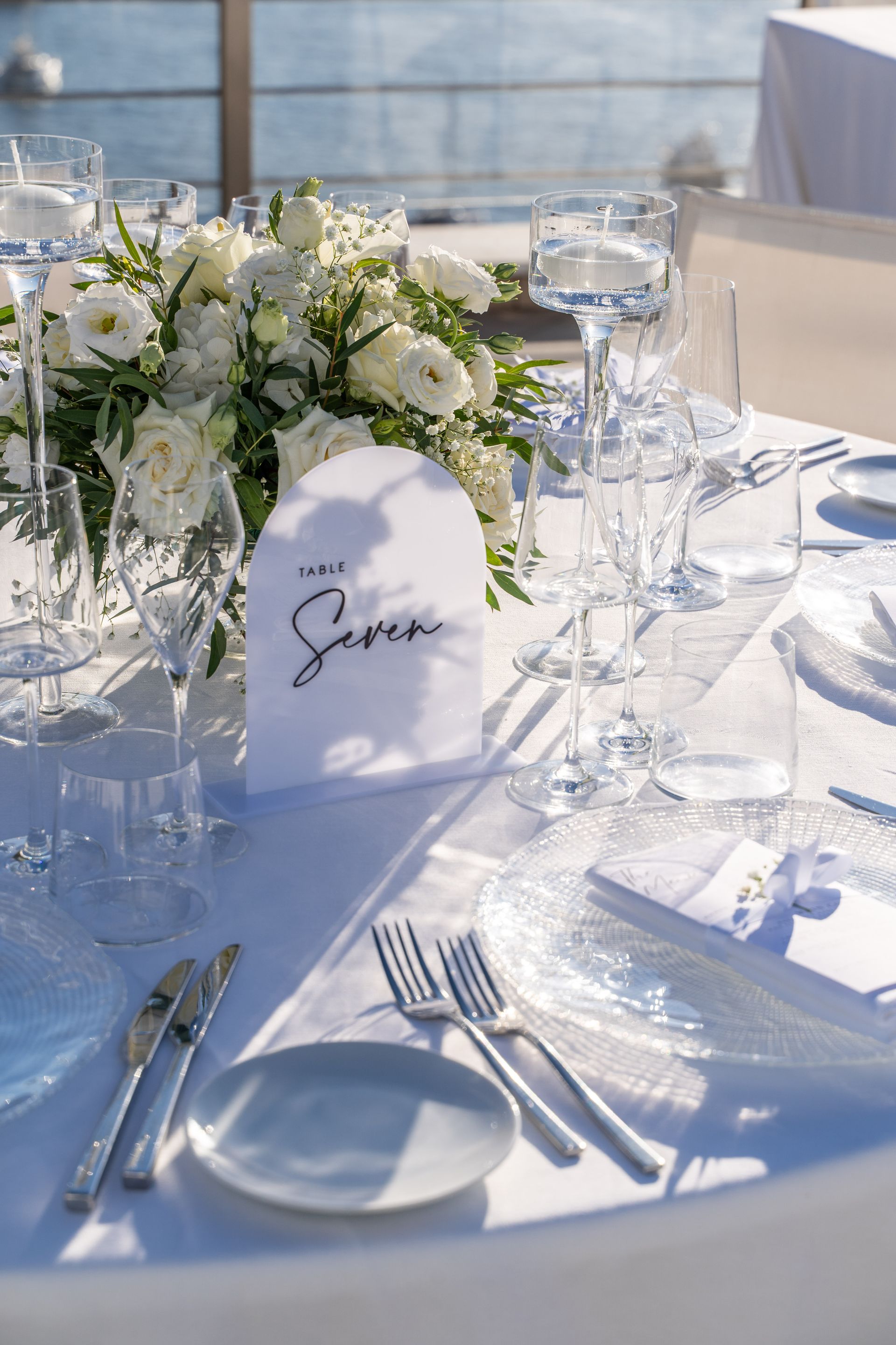 An elegant wedding table setting outdoors by the water with white floral centerpieces, crystal glasses, and a sign.