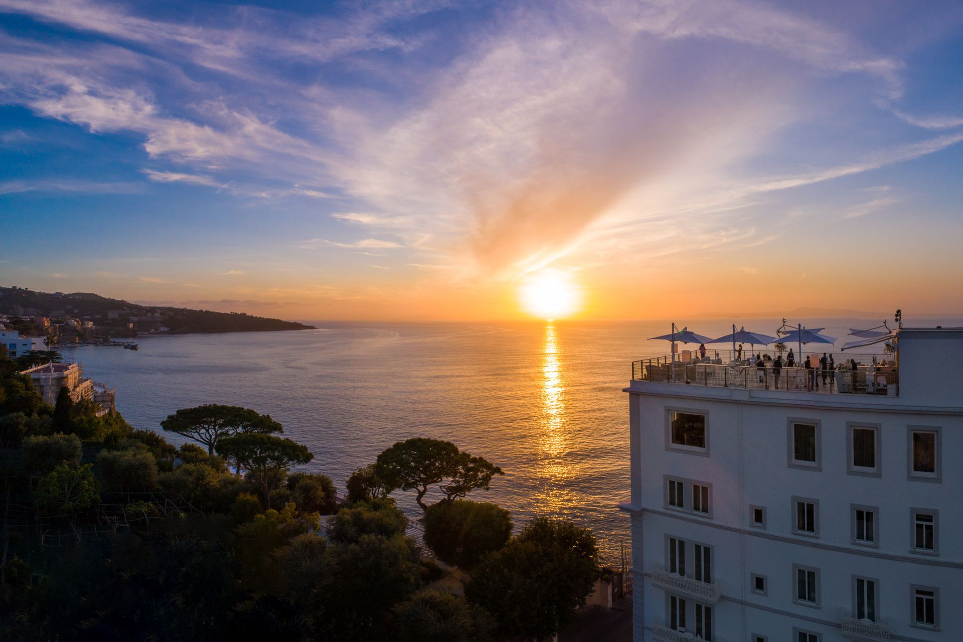 A sunset over the ocean, featuring a white building with an open-air rooftop terrace overlooking the calm, sparkling sea.