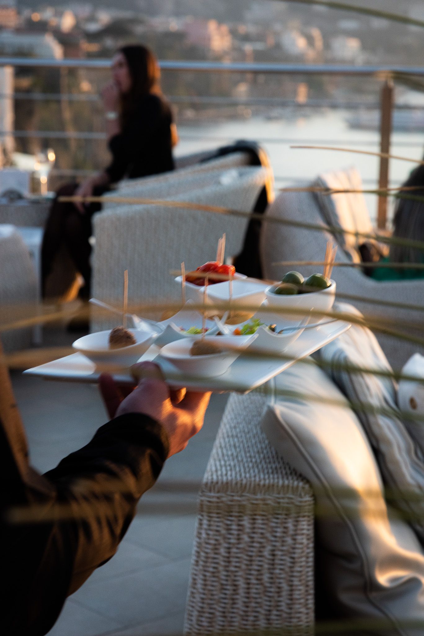 In un locale panoramico sul tetto, al tramonto, un cameriere tiene in mano un vassoio con delle piccole ciotole di antipasti, mentre sullo sfondo si intravede una persona sfocata seduta.