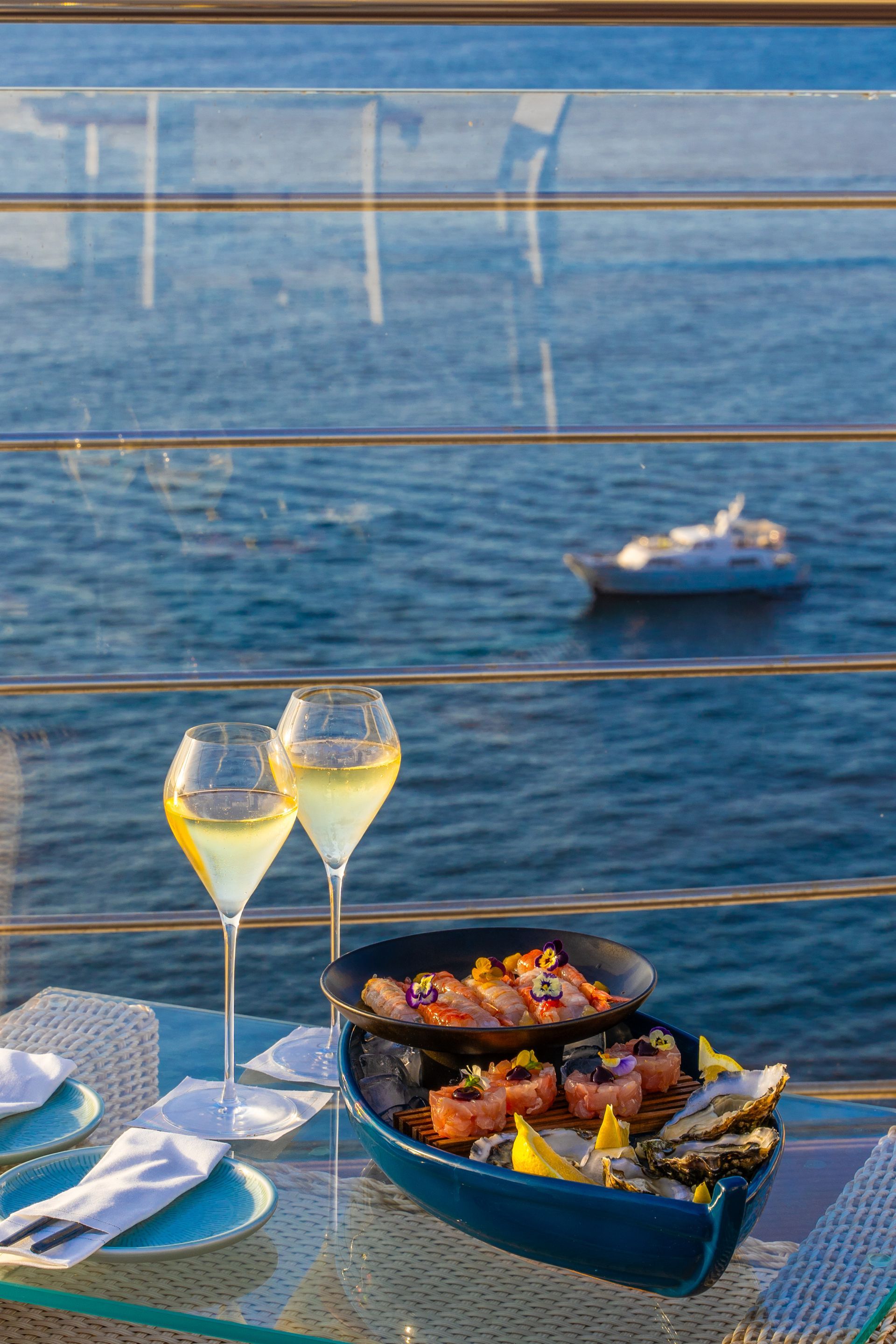 Due bicchieri di vino bianco accanto a un piatto di frutti di mare a forma di barca, su un tavolo di vetro con vista sul mare blu e su una barca.