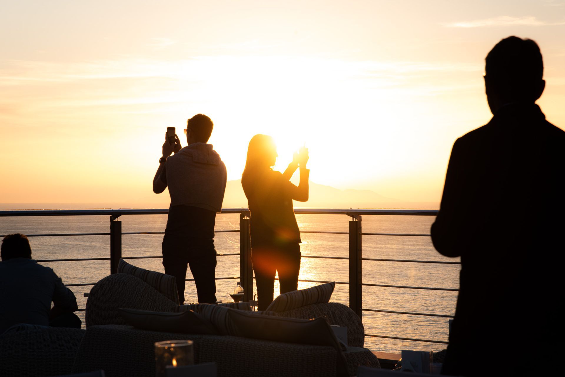 Sagome di persone su un balcone affacciato sull'oceano, intente a immortalare un tramonto con i loro smartphone.