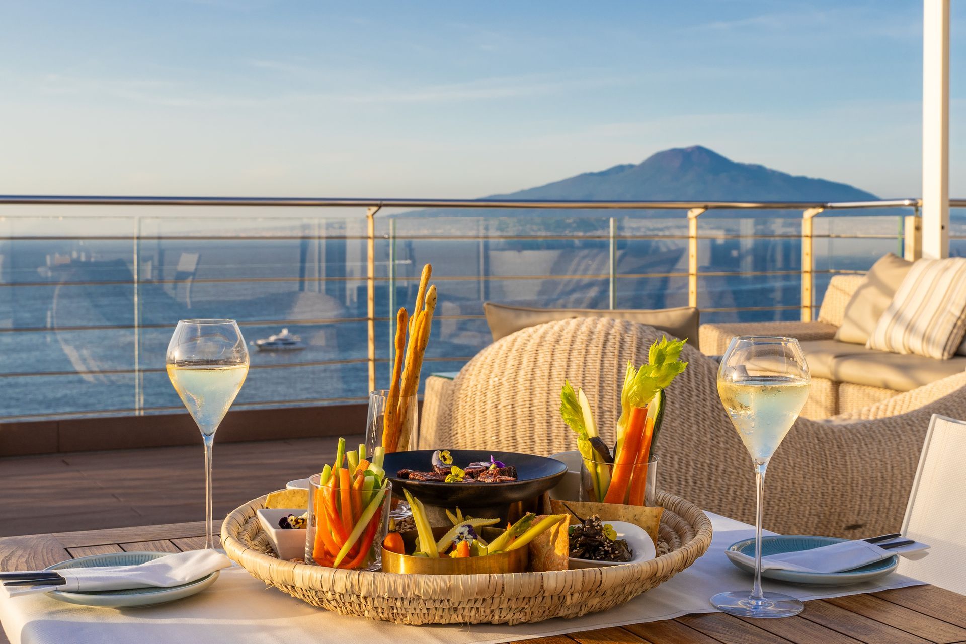 Piatto di antipasti e calici di vino su una terrazza con vista sul mare e sul Vesuvio al tramonto.
