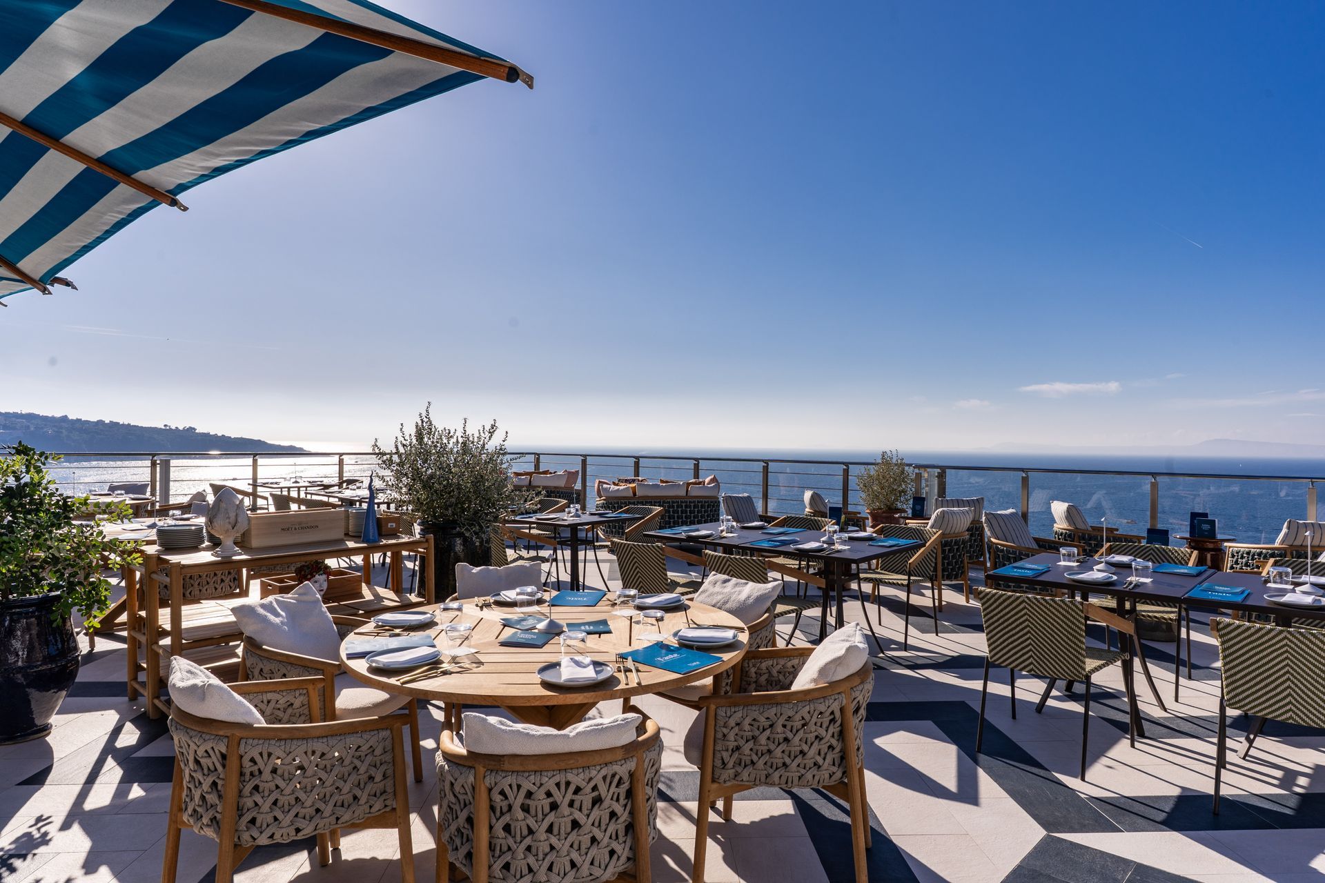 Mobili da salotto in vimini su una terrazza soleggiata con vista sull'oceano, con grandi ombrelloni bianchi, piante in vaso e lanterne decorative.