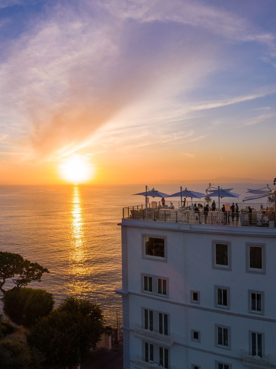 A sunset illuminates the ocean next to a white multi-story hotel with people on the rooftop terrace.