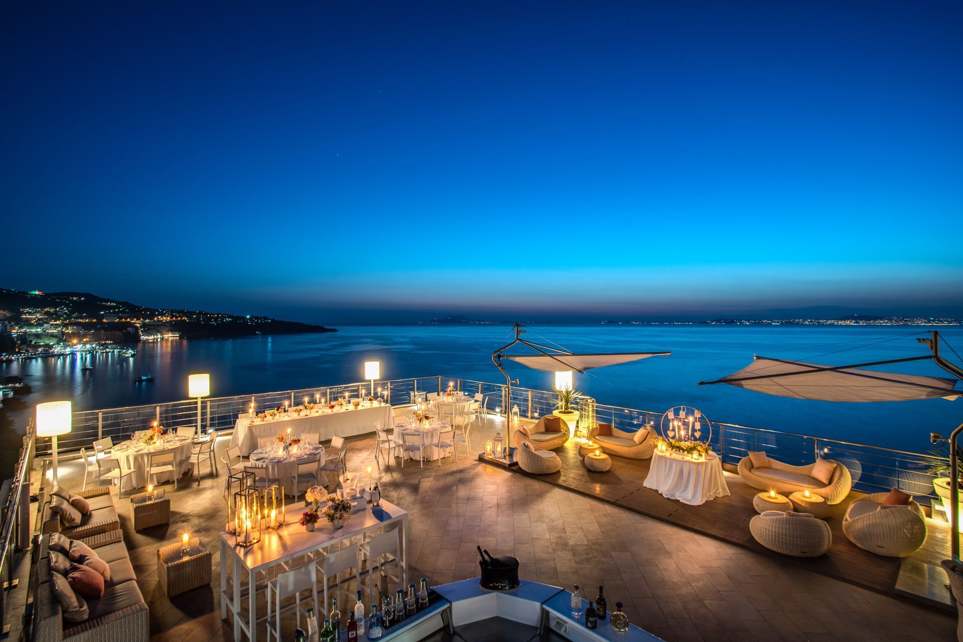 A high-angle view of a seaside restaurant terrace at twilight, featuring elegant dining tables and lounge seating.