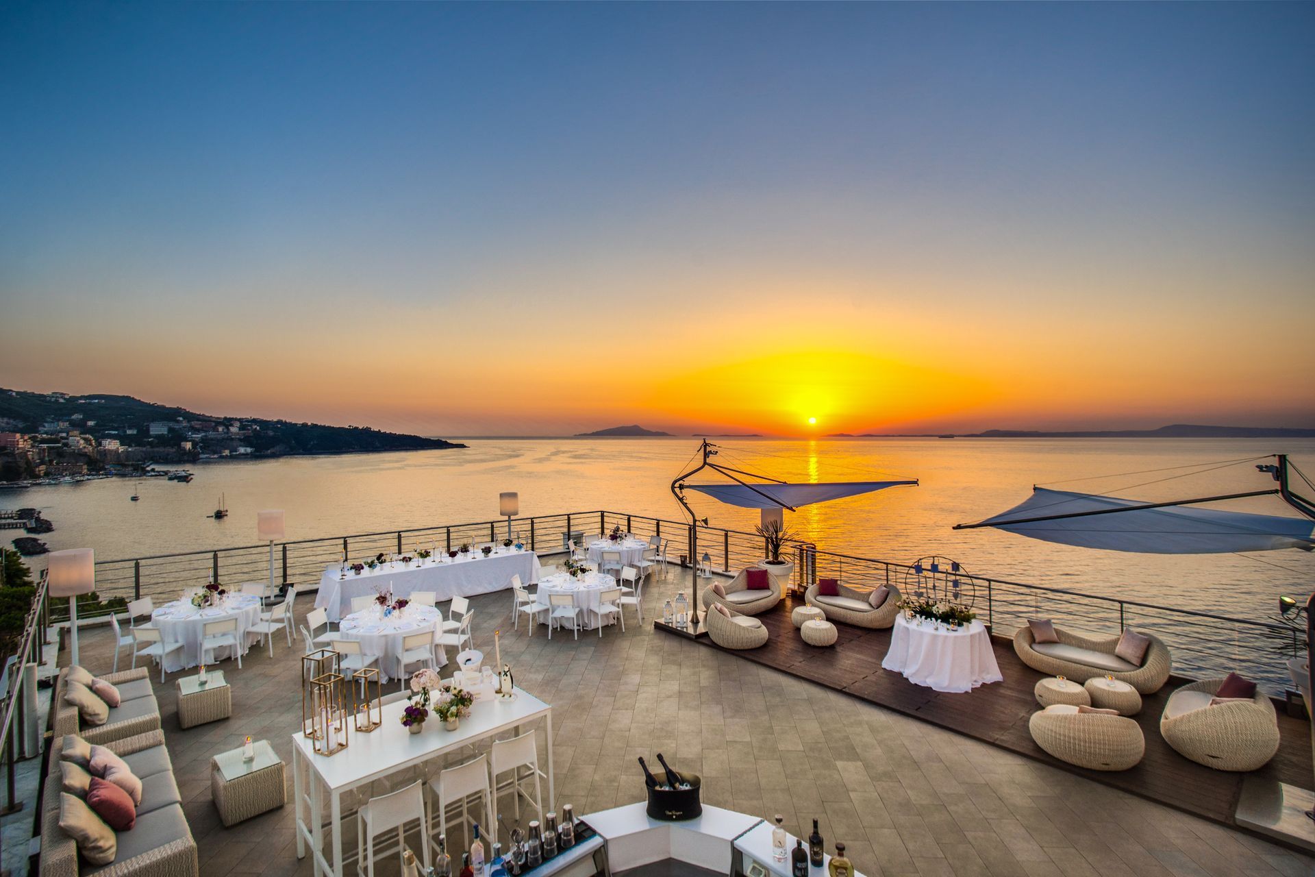A high-angle view of a seaside terrace set for an event at sunset, with tables, seating, and panoramic ocean views.