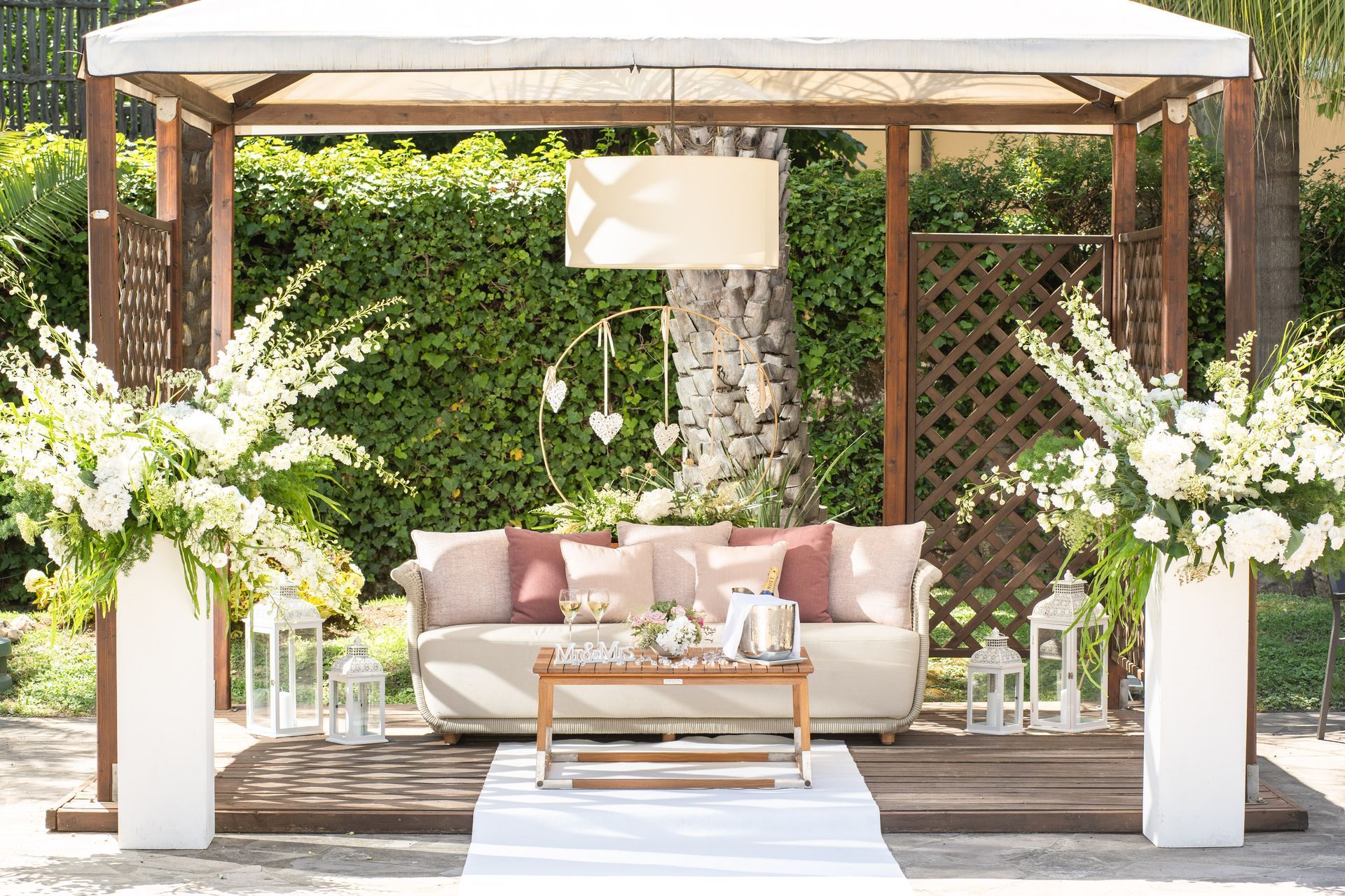 A gazebo set up for an event with a white sofa, coffee table, and large floral arrangements on pedestals.