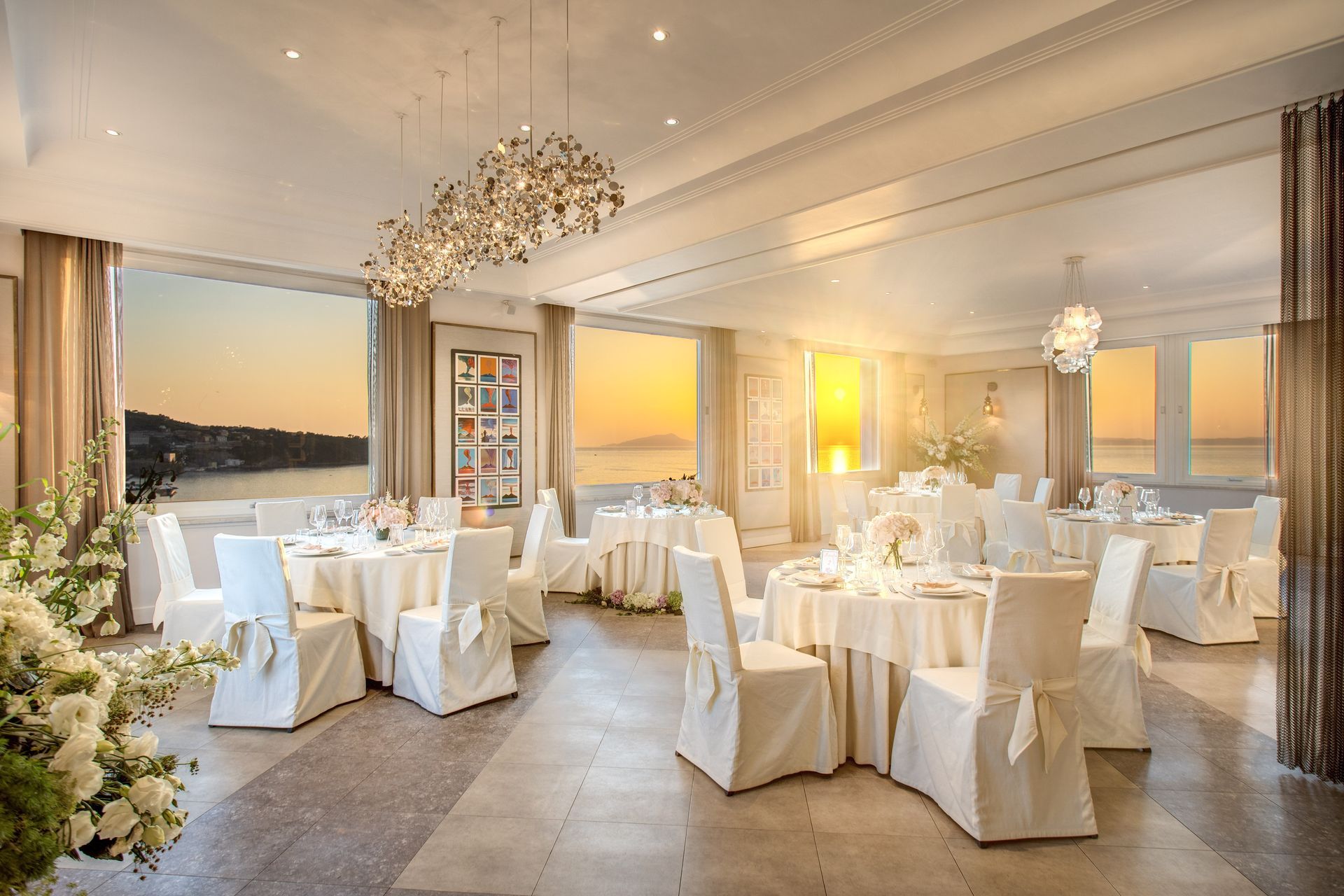 Elegant dining room with white-clothed tables, chairs, and chandeliers, overlooking a sunset sea view.