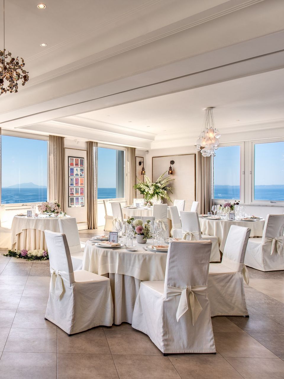 Elegant, sunlit dining room with white-clothed round tables, chairs, and large windows overlooking the ocean.