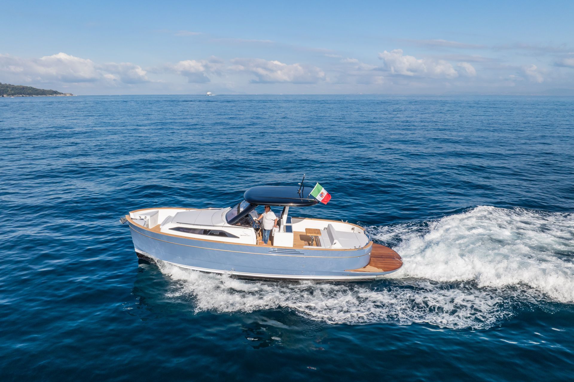 A light blue motorboat with a dark canopy and an Italian flag cruises on the open, deep blue sea.