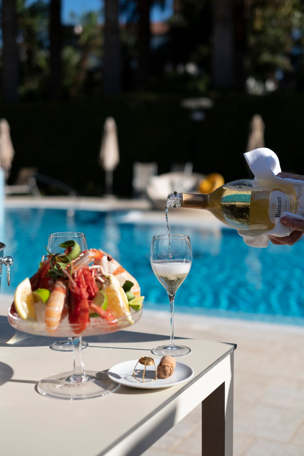 A hand pours white wine into a glass next to a seafood platter on a table beside a swimming pool.