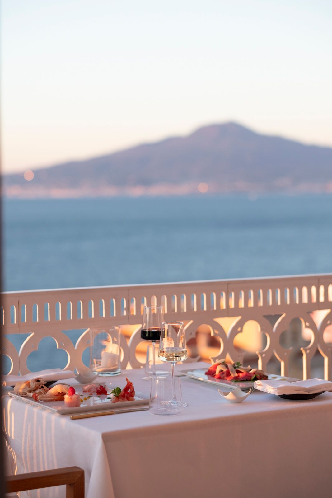 A table set for dinner on a balcony overlooking the sea and Mount Vesuvius at sunset.