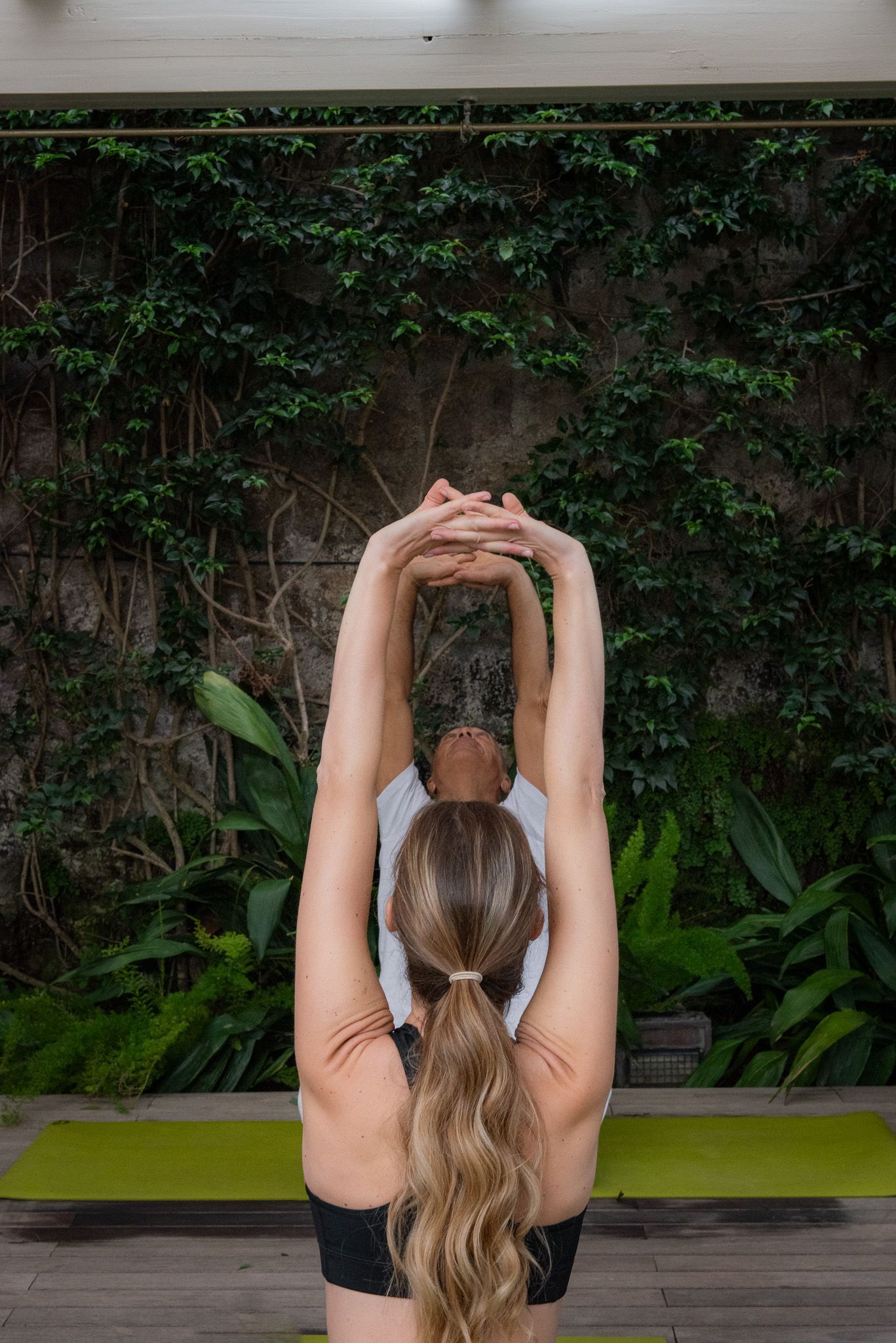 Una persona con i capelli raccolti esegue una posizione yoga, allungando le braccia verso l'alto con le mani giunte davanti a un muro verdeggiante.