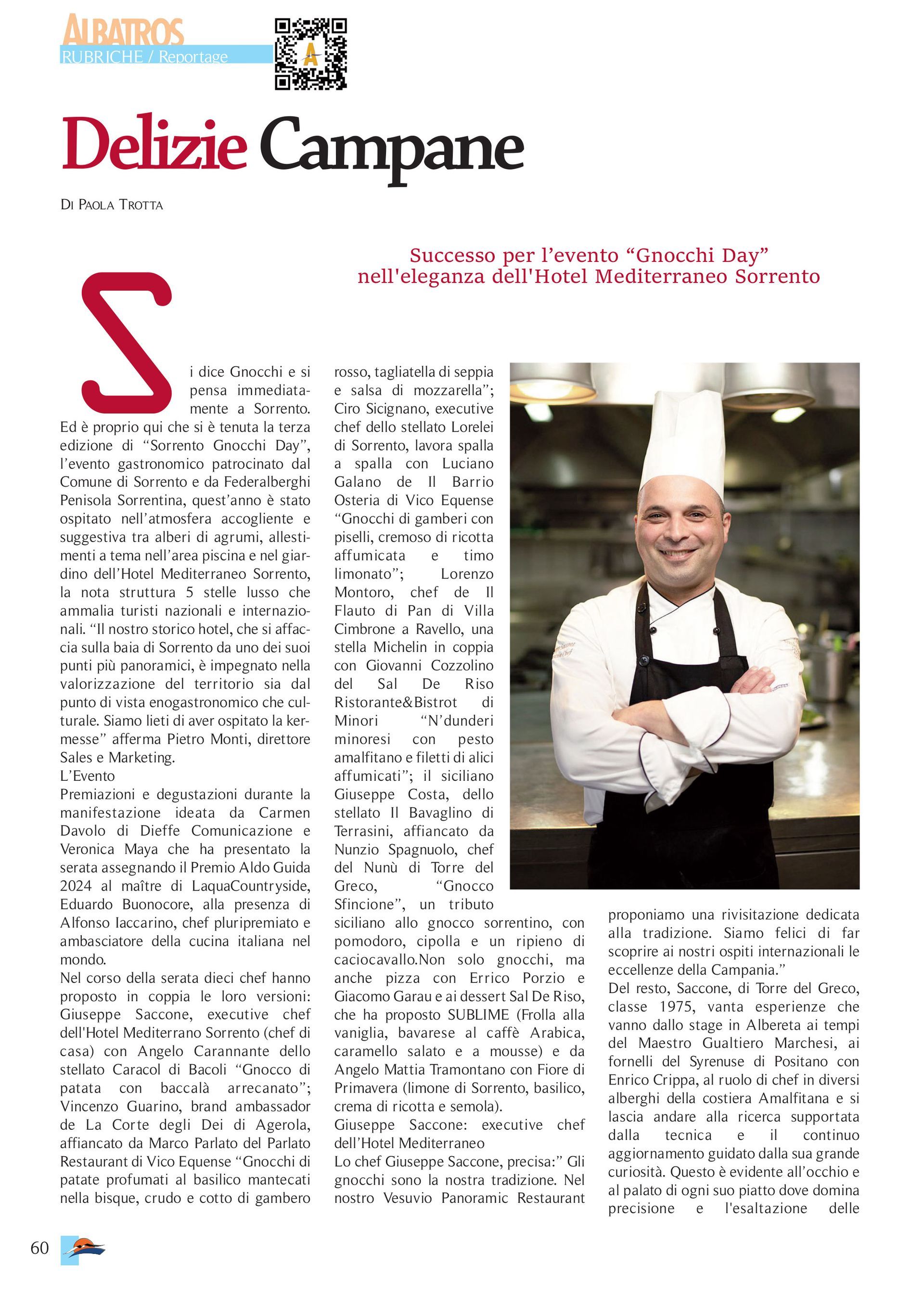 A smiling chef in a white uniform and black apron stands with arms crossed in a professional kitchen.