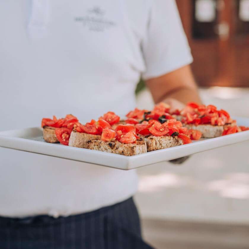 Una persona in uniforme bianca tiene in mano un vassoio rettangolare di bruschette guarnite con pomodori a cubetti.