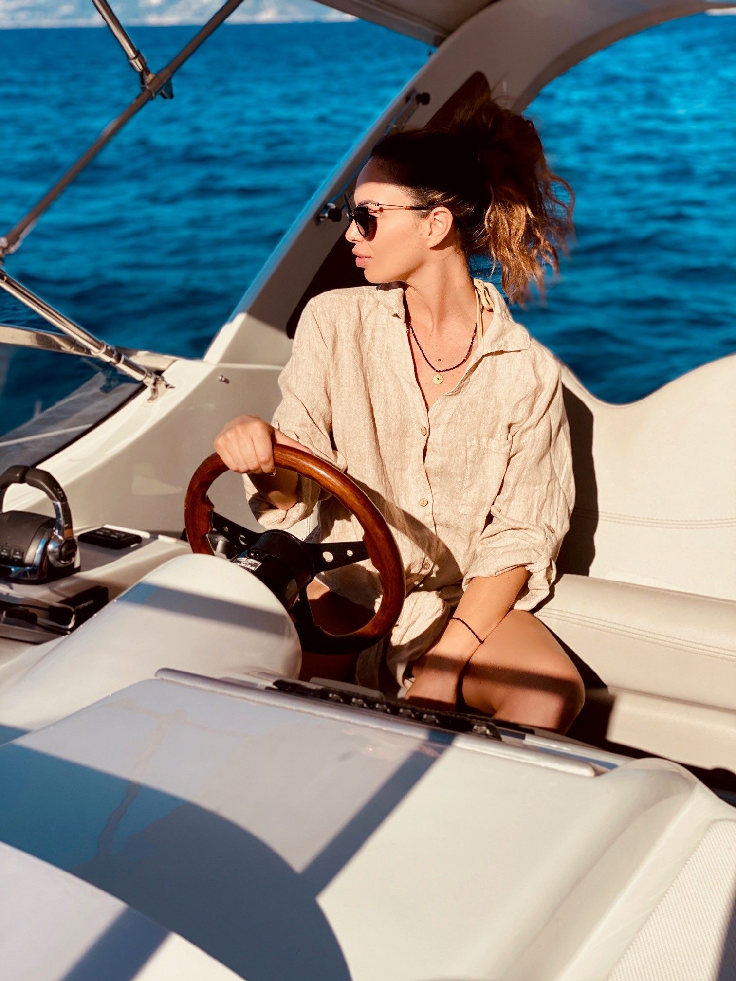 A person in a beige shirt and sunglasses steers a boat on blue water, looking toward the horizon.