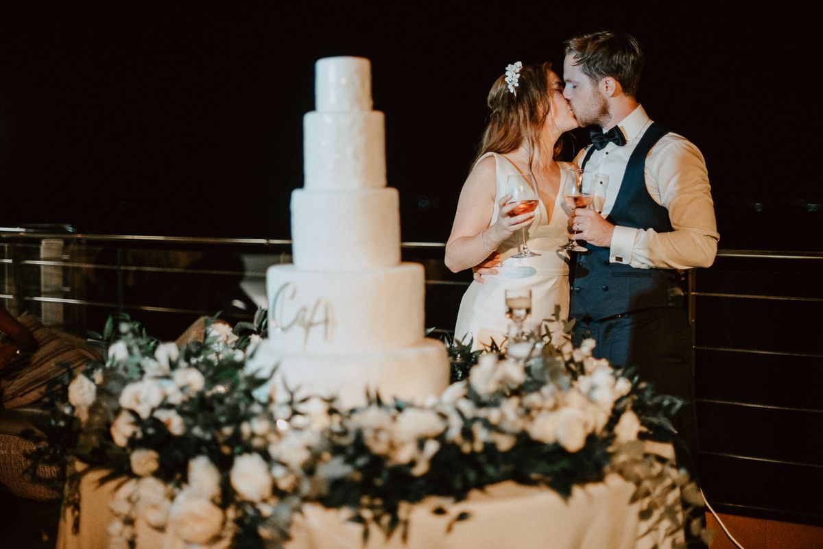 A couple kisses beside a tall, five-tiered white wedding cake decorated with flowers at an evening outdoor reception.