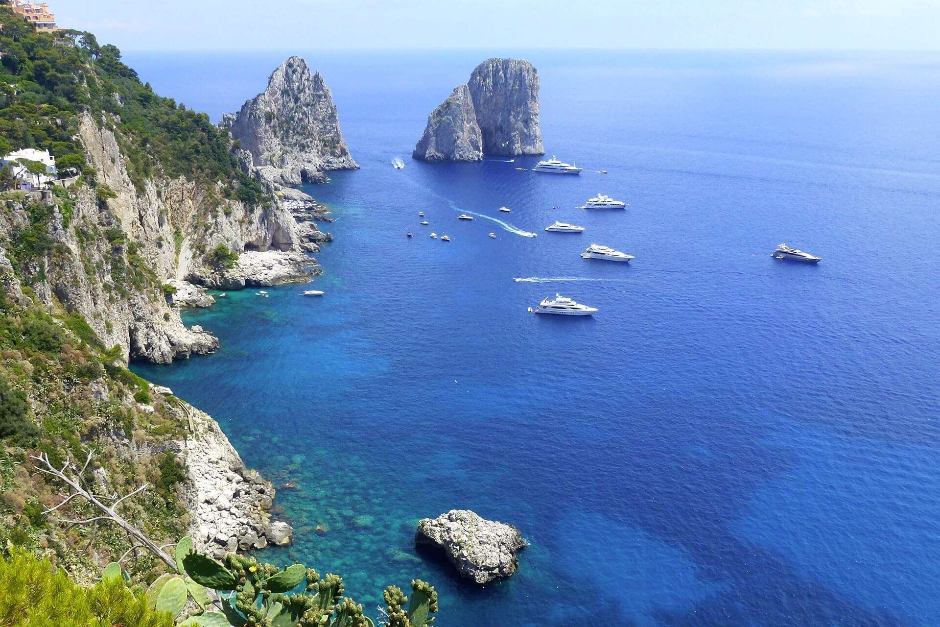 Una vista panoramica dei Faraglioni di Capri che emergono dalle acque blu intenso del Mediterraneo, con diverse imbarcazioni nelle vicinanze.