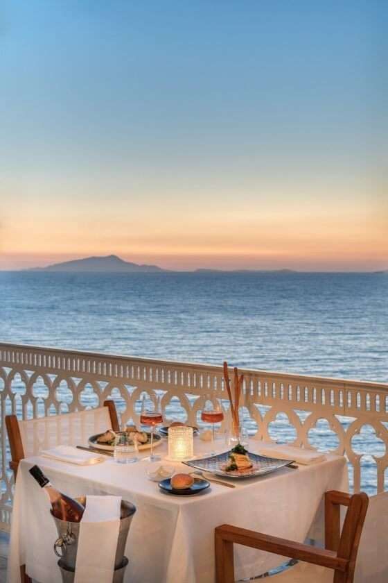 Una cena romantica per due su un balcone con vista sull'oceano al tramonto, con una bottiglia di vino in fresco in un secchiello.