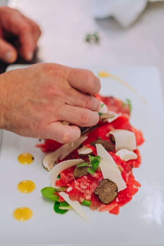 La mano di uno chef guarnisce un piatto di carpaccio di manzo con sottili scaglie di formaggio, erbe aromatiche e fette di tartufo nero su un piatto bianco.