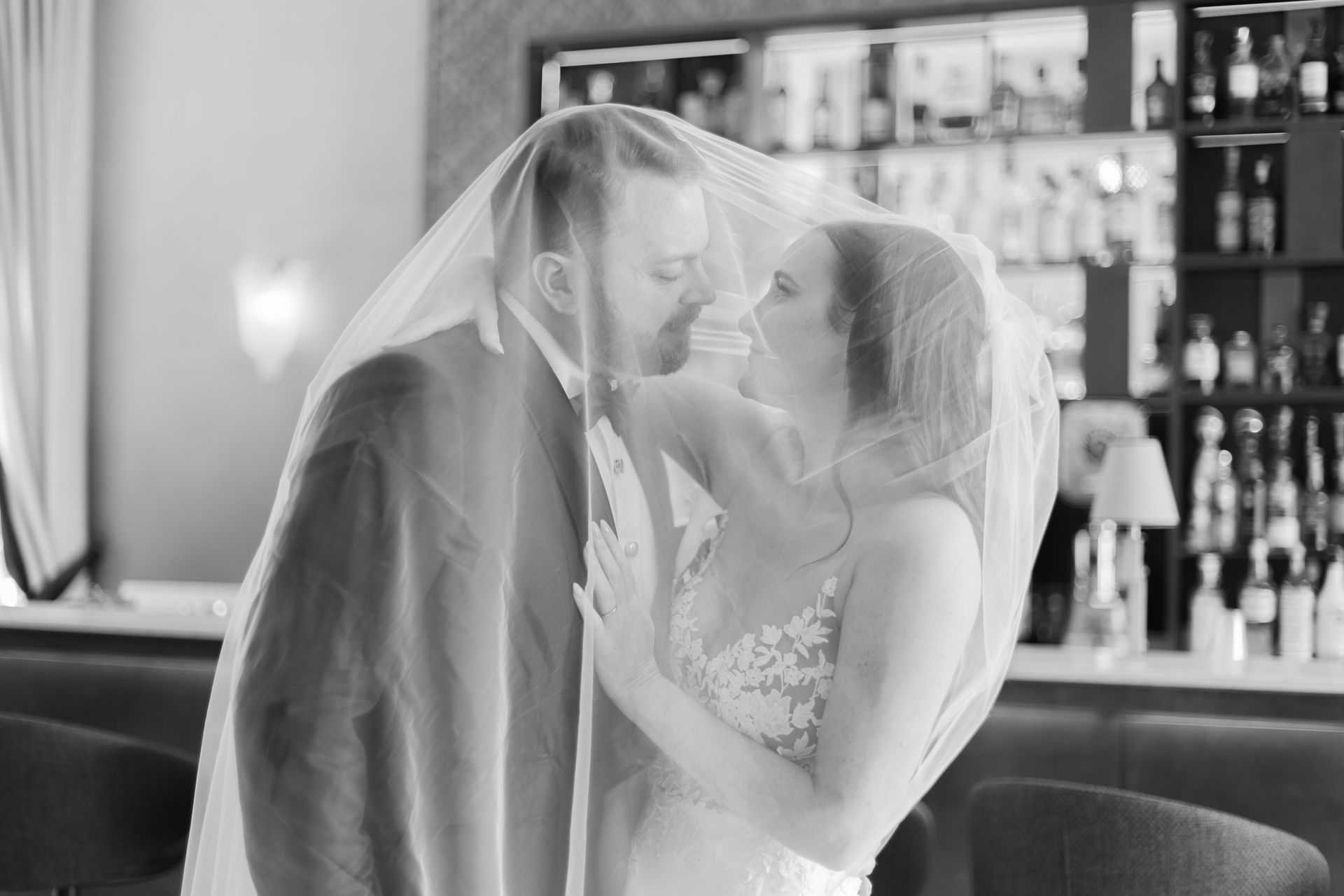 Una foto in bianco e nero di una coppia che condivide un momento intimo sotto un velo da sposa davanti a un bar.