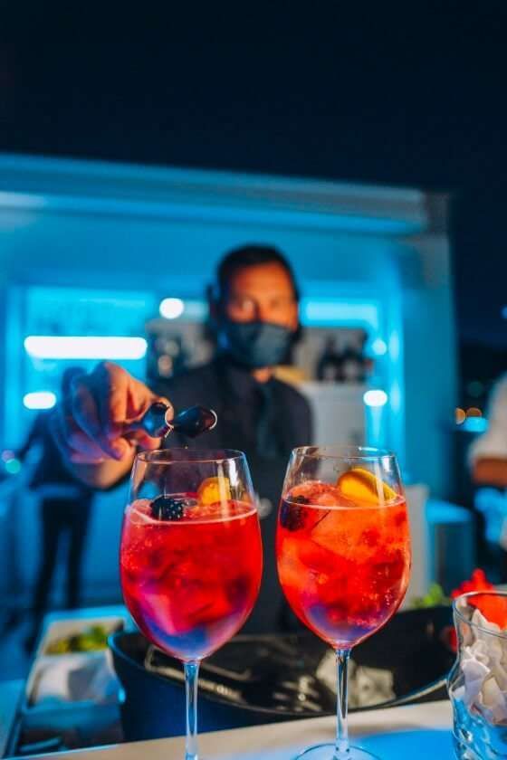 Un barista con la mascherina posiziona una guarnizione in uno dei due cocktail rosso acceso serviti in bicchieri sul bancone.