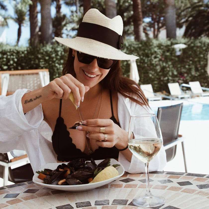 Una persona con un cappello fedora e occhiali da sole sorride mentre sprema uno spicchio di limone su una ciotola di cozze a bordo piscina.