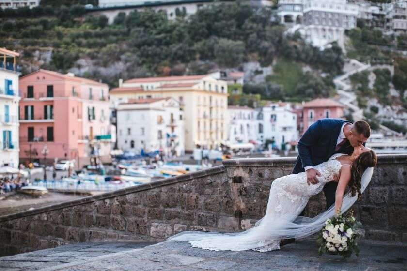 A bride and groom in wedding attire embrace in a dip while kissing on a stone terrace overlooking a coastal Italian town.