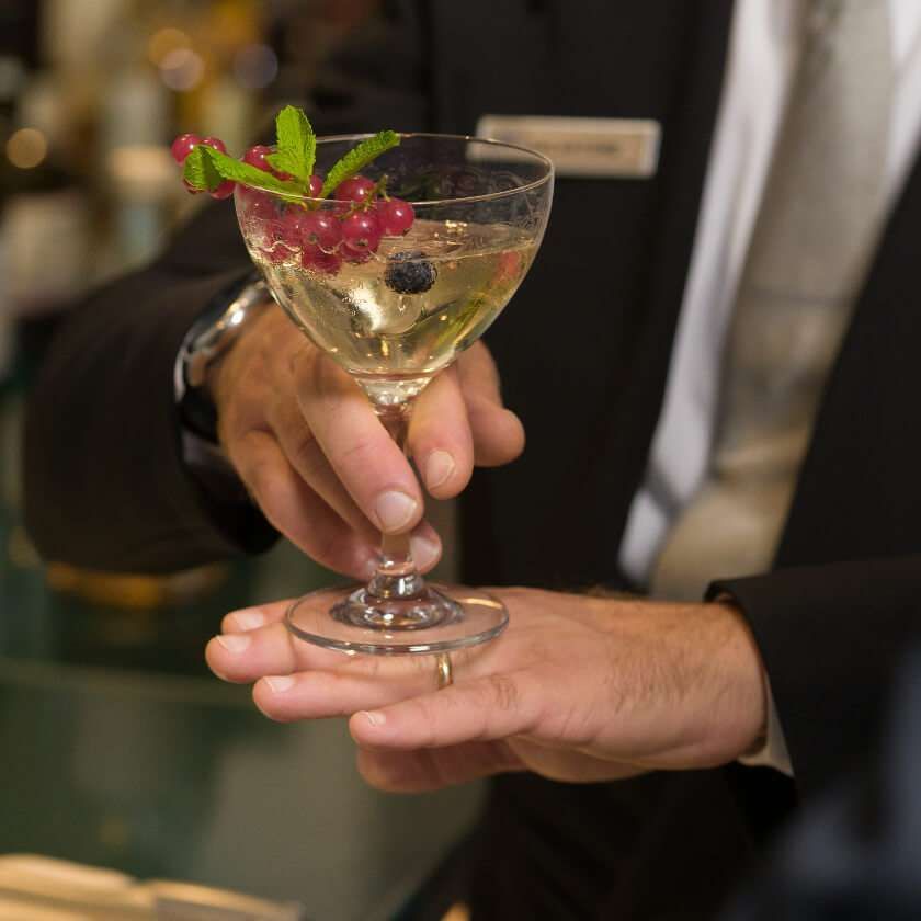 Un uomo in giacca e cravatta serve un cocktail in un bicchiere con stelo, guarnito con menta e ribes rosso.