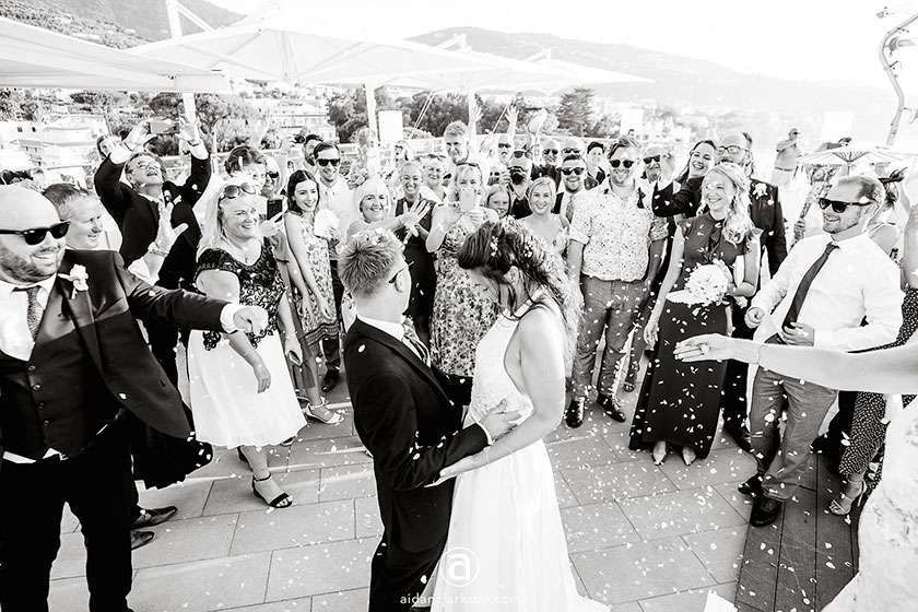 A wedding couple stands together in a crowd as guests toss confetti over them during an outdoor reception.