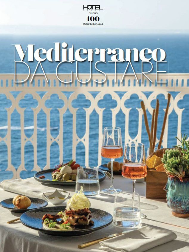 A dining table set for two on a balcony overlooking the sea, featuring plates of food, glasses of wine, and breadsticks.